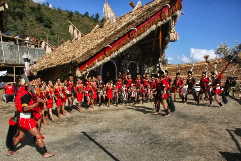 Naga dancers in red shawls performing in front of a thatched morung at the Hornbill Festival